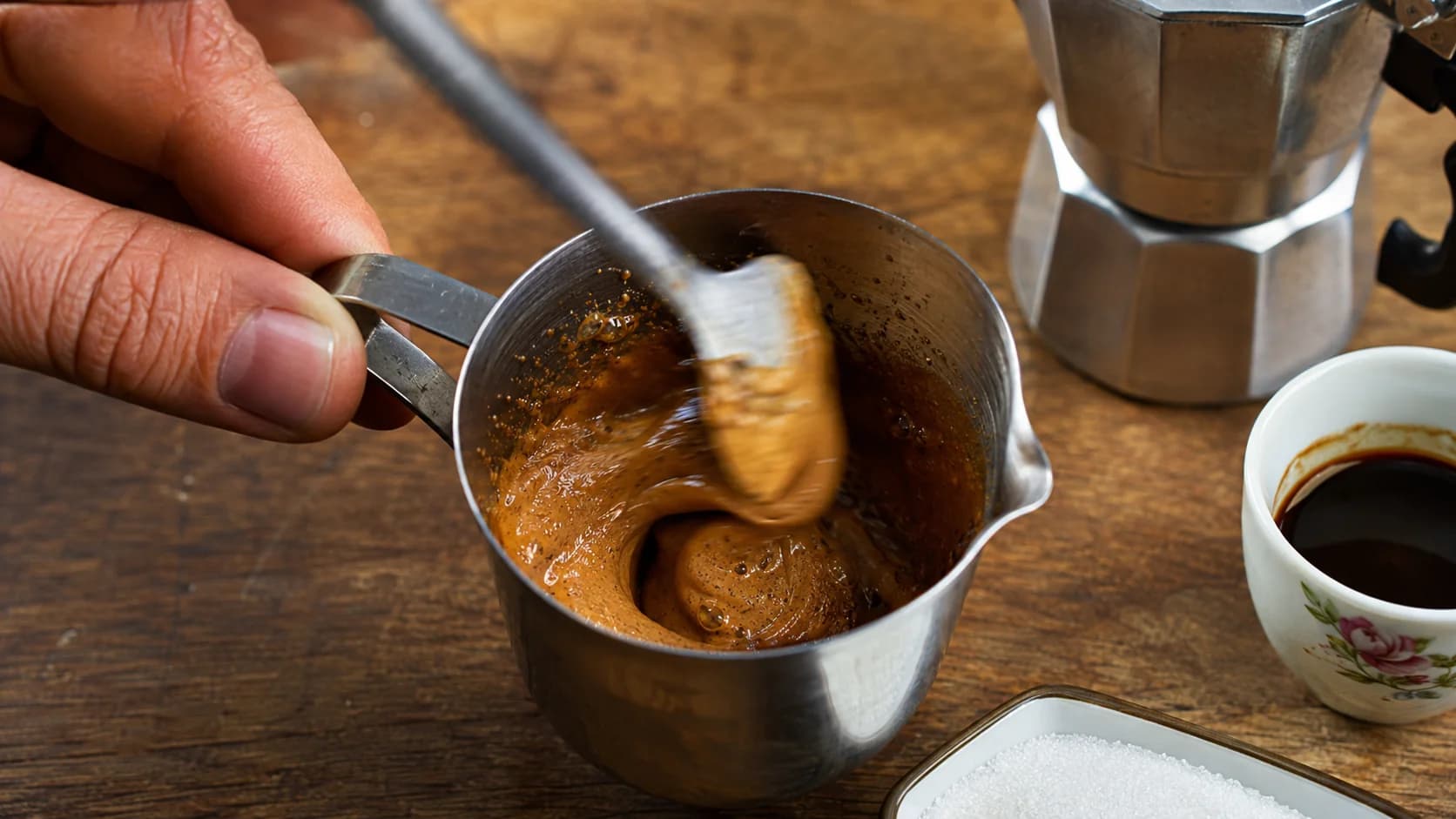 Cafecito espumita being whipped in a metal cup with moka pot coffee and sugar nearby