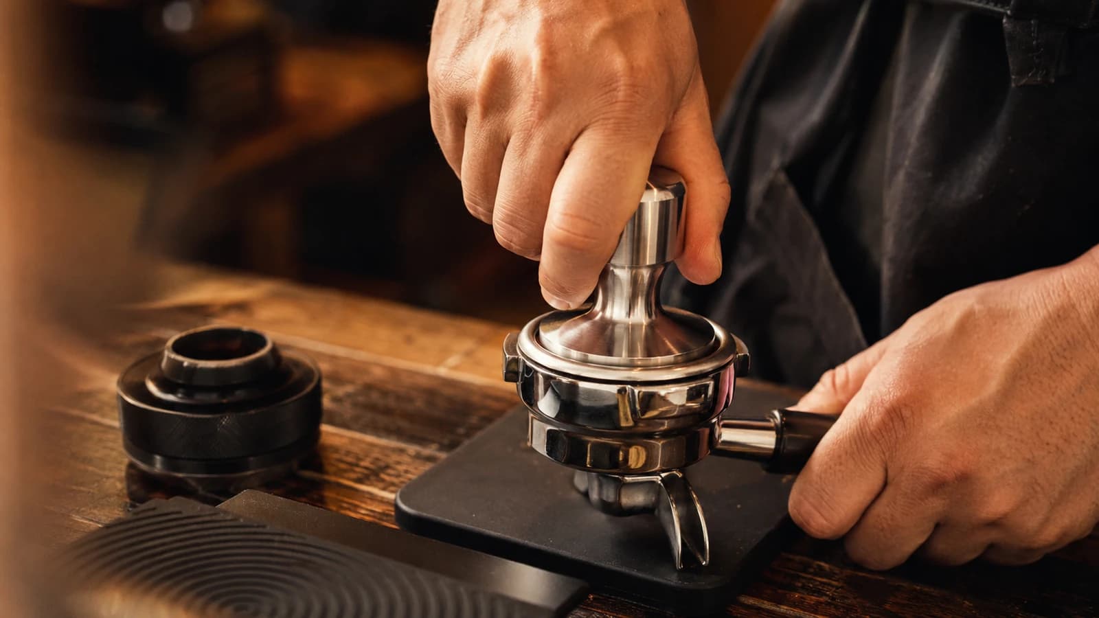Barista using a metal tamper to compress coffee evenly in an espresso portafilter on a tamping mat.