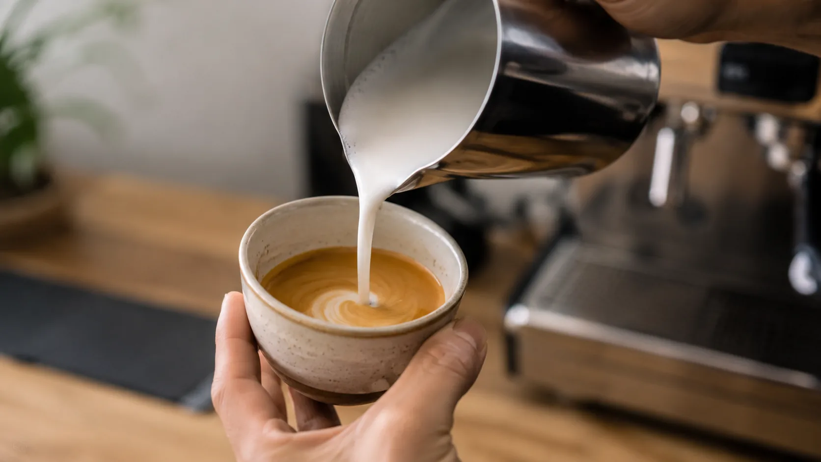 Steamed microfoam being poured into a flat white beside an espresso machine