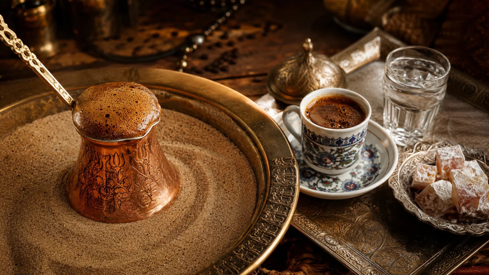 Turkish coffee foaming in a copper cezve set in hot sand beside a served cup